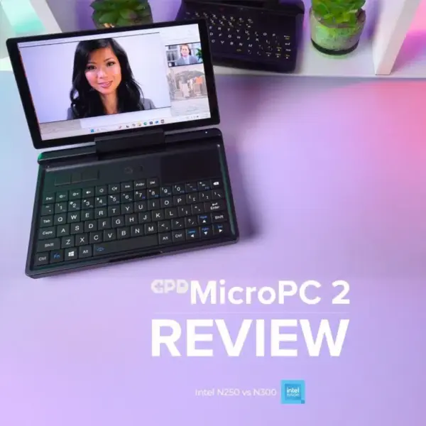 A thumbnail for a video review of the GPD MicroPC 2 mini-laptop. The black device is open and angled on a desk with a pink and purple gradient surface. The screen displays a video conference call featuring a woman's face. In the background, there is a white shelf with potted plants and another GPD MicroPC 2. Large white text overlaid on the desk reads "GPD MicroPC 2 REVIEW," with smaller text below it saying "Intel N250 vs N300" next to the Intel inside logo.