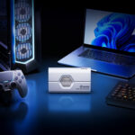 A lifestyle shot of the white SSD reader placed centrally on a blue-lit desk setup, flanked by a gaming PC tower, a game controller, and a laptop.