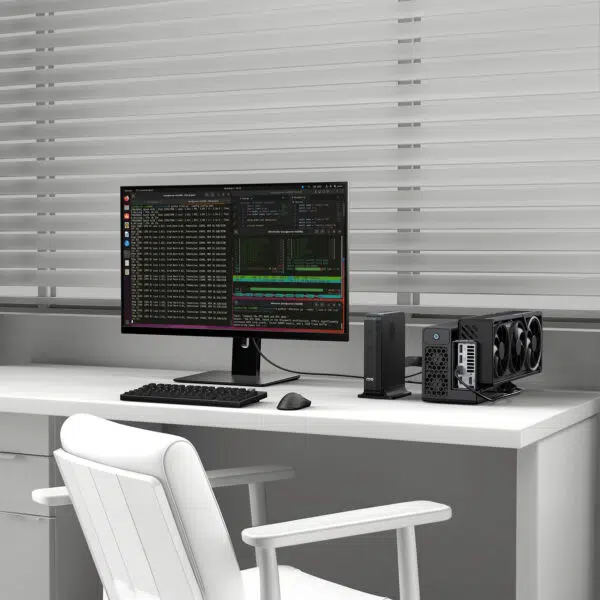 Configurazione della scrivania con un monitor widescreen che visualizza codice e finestre del terminale, un PC compatto e una GPU esterna sulla destra, una sedia bianca e tende sullo sfondo.